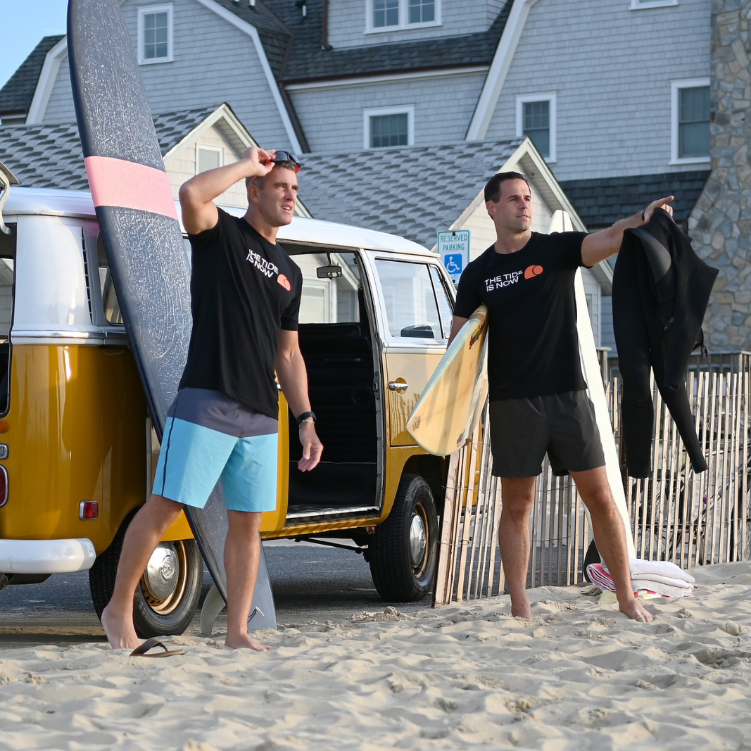 Two men on the beach by a VW van with surfboards with The Tide Is Now tshirts on