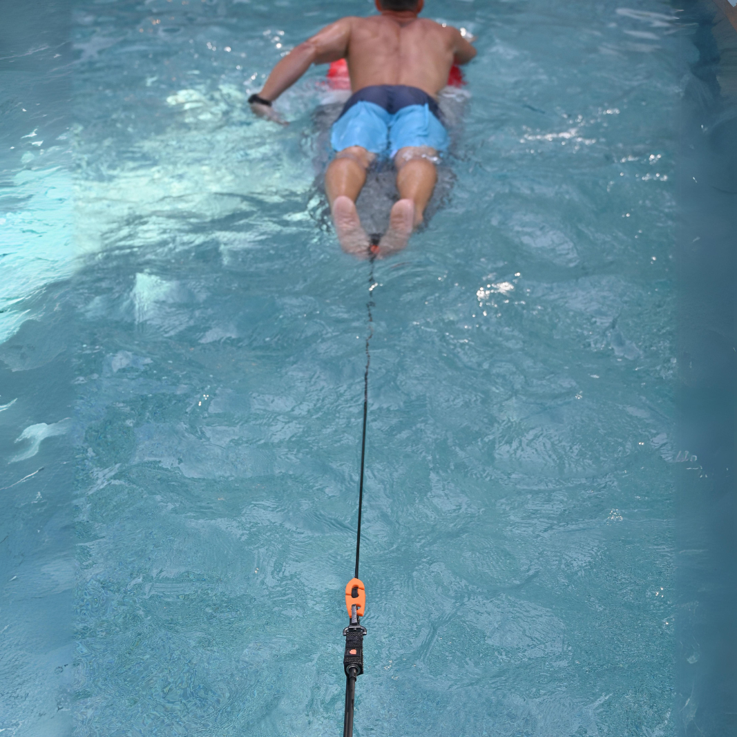 A view of a man on a surfboard in a swimming pool tethered to the edge with the 365Waves Surf Resistance Trainer