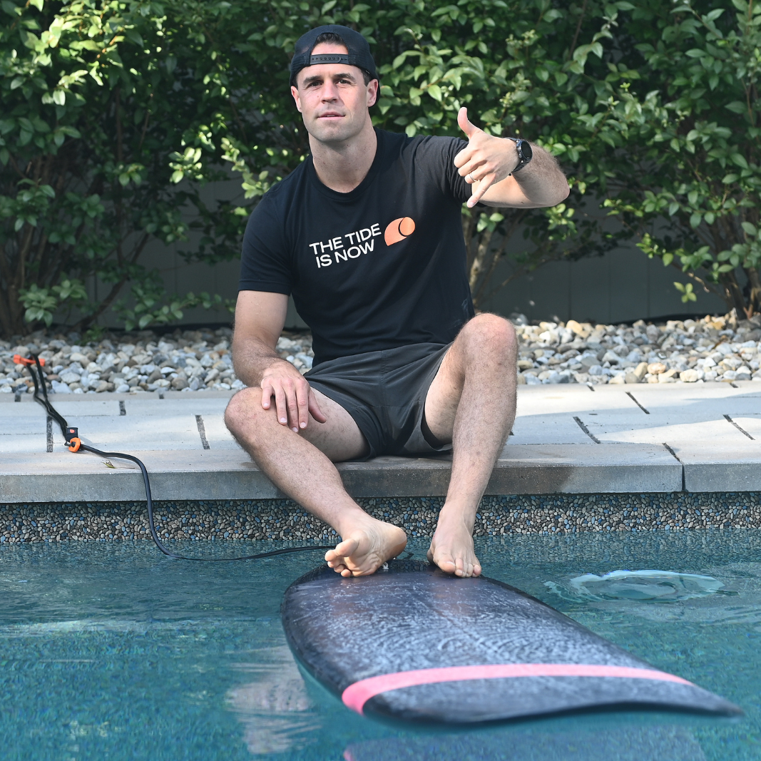 365Waves founder sitting on the edge of a swimming pool wearing a black The Tide Is Now tshirt