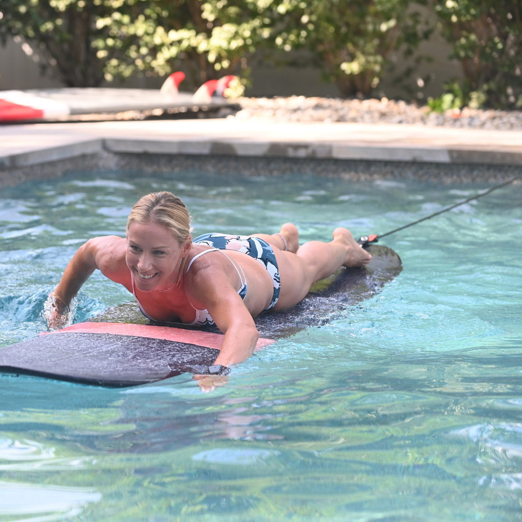A woman in a swimming pool paddling on a surfboard that is tethered to the edge with the 365Waves Surf Resistance Trainer